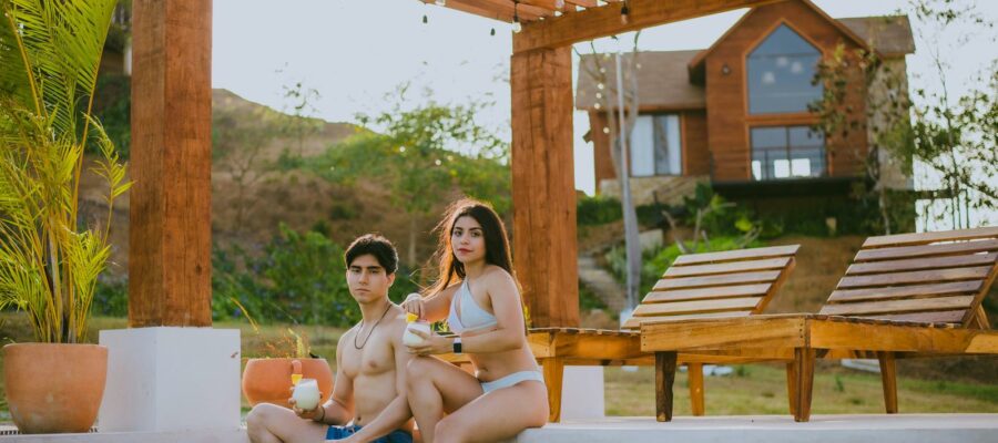 Young couple enjoying drinks by the poolside under a rustic pergola on a sunny day.