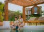 Young couple enjoying drinks by the poolside under a rustic pergola on a sunny day.